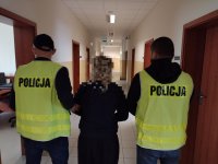 Zdjęcie kolorowe. Dwóch policjantów w kamizelkach odblaskowych z napisem POLICJA prowadzi kobietę przez korytarz