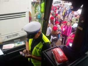 Zdjęcie kolorowe. Policjant dokonuje kontroli autokaru. Na zewnątrz oczekuje grupa dzieci