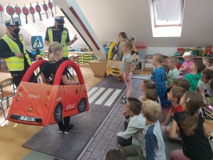 Zdjęcie kolorowe. Policjantka z elementami miasteczka ruchu drogowego bawi się z dziećmi aranżując sytuacje mogące wystąpić na drodze