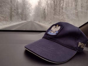 Zdjęcie kolorowe. Granatowa czapka policyjna lezy na podszybiu. W tle widać zaśnieżone pobocza, drzewa i drogę