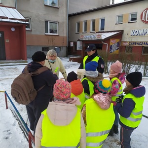 Zdjęcie kolorowe. Policjantka i dzieci ubrane w kamizelki odblaskowe rozmawiają z seniorami na ulicach Szczecinka