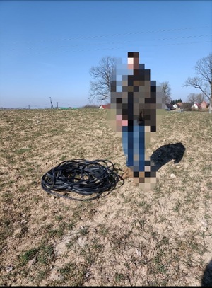 Zdjęcie kolorowe. Na polu stoi mężczyzna przy czarnym zwoju kabla