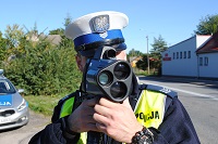 Zdjęcie kolorowe. Policjant ruchu drogowego w białej czapce i kamizelce odblaskowej dokonuje pomiaru prędkości poruszających się po drodze pojazdów urządzeniem pomiarowo kontrolnym