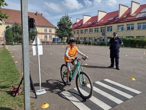 Zdjęcie kolorowe. Uczeń w kasku i kamizelce na rowerze pokonuje tor. W oddali jego jeździe przygląda się policjant.
