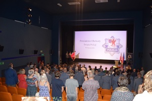 Zdjęcie kolorowe. Uczestnicy uroczystości stojąc na baczność śpiewają hymn. W tle na scenie widać poczet sztandarowy oraz prowadzącego uroczystość lektora.