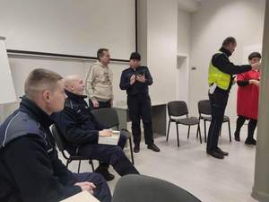 Zdjęcie kolorowe. Policjanci podczas spotkania z osobami niesłyszącymi. Wszyscy siedzą przy stołach, które tworzą koło. W centralnym miejscu siedzą umundurowani policjanci.