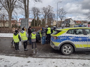Zdjęcie kolorowe. Wspólne działania uczniów i policjantów ruchu drogowego z okazji Dnia Kobiet. Ubrani w kamizelki odblaskowe uczniowie stoją u boku policjantów na tle zatrzymanego do kontroli samochodu.