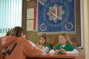 Zdjęcie kolorowe. Uczniowie siedzą przy stoliku i piszą test. Za ich plecami na ścianie w szklanej gablocie wisi sztandar KPP w Szczecinku