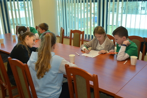 Zdjęcie kolorowe. Uczniowie siedząc przy stolikach rozwiązują test