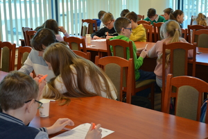 Zdjęcie kolorowe. Uczniowie siedząc przy stolikach rozwiązują test