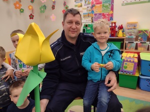 Zdjęcie kolorowe. Policjant w ręku trzyma papierowego kwiata wykonanego przez dzieci. Na kolanie policjanta siedzi chłopiec w niebieskiej bluzie.