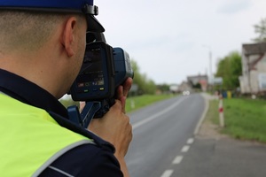 Zdjęcie kolorowe. Na pierwszym planie policjant ruchu drogowego dokonuje pomiaru prędkości