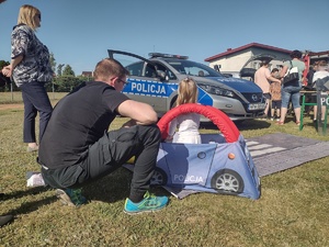 Zdjęcie kolorowe. Malutkie dziecko siedzi w piankowym samochodziku z napisem policja. Obok kuca mężczyzna. W tle widać policyjny radiowóz