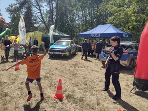Zdjęcie kolorowe. Policjantka przygląda się jak chłopiec w pomarańczowej koszulce i alkogooglach pokonuje slalom pomiędzy ustawionymi w szeregu pachołkami drogowymi