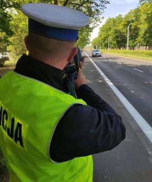 Zdjęcie kolorowe. Policjant ruchu drogowego odwrócony plecami dokonuje pomiaru prędkości