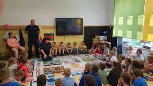 Zdjęcie kolorowe. Wokół planszy wielkoformatowej gry rozłożonej w szkolnej klasie siedzą dzieci oraz umundurowani policjanci