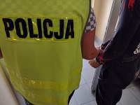 Zdjęcie kolorowe. Duże zbliżenie policjanta w kamizelce odblaskowej z napisem policja, odwróconego plecami. Z prawej strony stoi mężczyzna z założonymi kajdankami