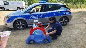 Zdjęcie kolorowe. Na tle radiowozu policjantka pomaga dziecku które siedzi w piankowym, niebieskim autochodziku z napisem policja