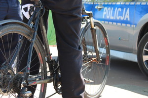 Zdjęcie kolorowe. Na pierwszym planie przez szprychy od koła do roweru widać policyjny radiowóz
