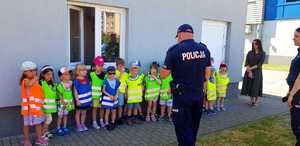 Zdjęcie kolorowe. Dzieci stoją w szeregu. Każde ma ubraną kamizelkę odblaskową przed nimi stoi policjant.