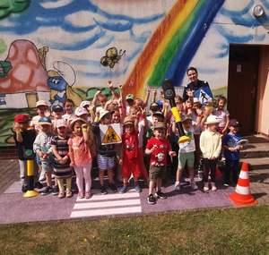 Zdjęcie kolorowe. Zdjęcie grupowe wszystkich uczestników spotkania z policjantką. Dzieci stoją na tle pomalowanej ściany budynku na której widać chmury i tęczę.