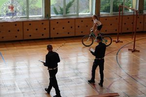 Zdjęcie kolorowe. Uczestnik jadąc na rowerze pokonują kolejną przeszkodę w tle policjant. Tor rozłożony jest na sali gimnastycznej.