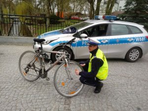 Na tle radiowozu policjant montuje w przednim kole roweru światełka odblaskowe