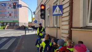 Zdjęcie kolorowe. Policjant przed przejściem dla pieszych z sygnalizacją świetlną omawia wszystkie znaki drogowe i zasady poruszania się na nim.