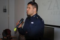 Zdjęcie kolorowe. Policjant trzyma mikrofon w dłoni. W tle białe tło ekranu