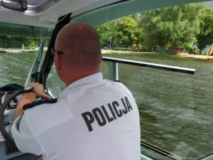 Zdjęcie kolorowe. Zdjęcie z kabiny łódki, na którym widać plecy policjanta w białej koszulce z napisem policja. Przez okna widać brzeg jeziora Trzesiecko