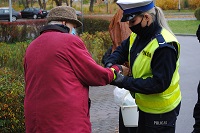 Zdjęcie kolorowe. Umundurowana policjantka z ruchu drogowego zakłada starszej kobiecie w czerwonej kurtce opaskę odblaskową na jej prawą rękę.