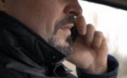 Zdjęcie kolorowe. Na zdjęciu wdać fragment twarzy mężczyzny siedzącego w samochodzie i rozmawiającego przez telefon.