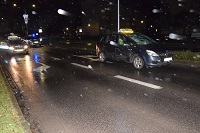 Zdjęcie kolorowe. Zdjęcie zrobione po zmroku, widać że pada deszcz. Na zdjęciu uchwycone miejsce zdarzenia na ul. JP II - ulica dwupasmowa w jednym kierunku. Z lewej strony na lewym i prawym pasie stoją policyjne radiowozy, z prawej strony stoi Opel z podświetlonym na dachu napisem TAXI