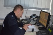 Zdjęcie kolorowe. Policjant siedzi przy biurku na którym stoi monitor komputera. Policjant rozmawia przez telefon