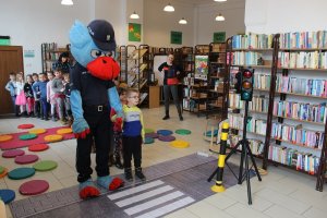 Zdjęcie kolorowe. W pomieszczeniu biblioteki policyjna maskotka Gryfuś przeprowadza dziecko przez matę z widocznym przejściem dla pieszych