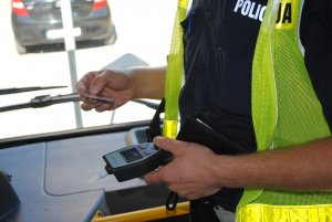 Zdjęcie kolorowe. Policjant trzyma w dłoni dokument tożsamości kierowcy oraz urządzenie do kontroli stanu trzeźwości