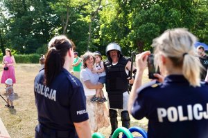 Zdjęcie kolorowe. Policjantki obserwują mężczyznę który pozuje do zdjęcia z rodziną w stroju wykorzystywanym w zwartych oddziałach prewencji podczas zabezpieczania imprez