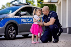 Zdjęcie kolorowe. Policjant na tle radiowozu rozmawia z małą dziewczynką w różowej sukience