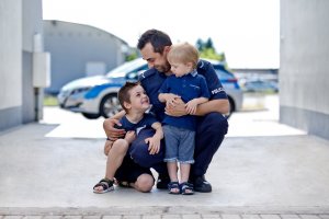 Zdjęcie kolorowe. Policjant pozuje z dwoma chłopcami na tle radiowozu.