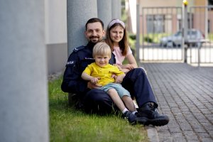 Zdjęcie kolorowe. Na trawie policjant siedzi w towarzystwie młodej dziewczynki i małego chłopca