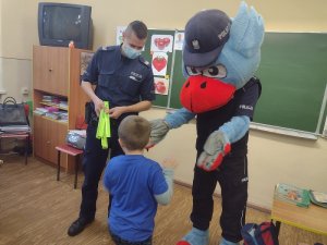 Zdjęcie kolorowe. Policjant wraz z Gryfusiem wręcza element odblaskowy.