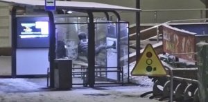 Zdjęcie kolorowe. Ujęcie z kamery monitoringu miejskiego. W centralnej części widać przystanek autobusowy w którym stoi mężczyzna w kapturze na głowie z podniesioną nogą. (kopiący ścianę przystanku)