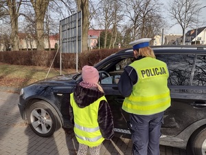 Zdjęcie kolorowe. Policjantka w obecności ucznia prowadzi kontrolę drogową zatrzymanego samochodu.