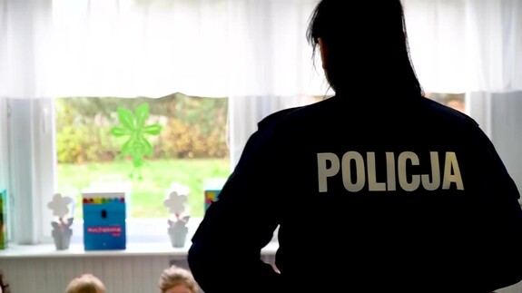 Zdjęcie kolorowe. Policjantka stoi odwrócona plecami. Na plecach widać biały napis policja