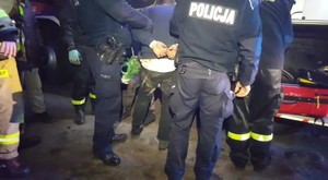 Zdjęcie kolorowe. Policjanci trzymają pod rękę zatrzymanego mężczyznę. Mężczyzna ma założone kajdanki. Wszyscy stoją odwróceni plecami.