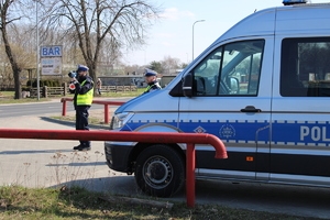 Zdjęcie kolorowe. Policjanci na drugim planie dokonują kontroli prędkości z jaką poruszają się samochody po drodze. Na pierwszym planie stoi oznakowany policyjny bus