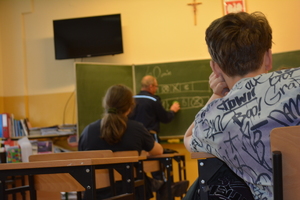 Zdjęcie kolorowe. Zdjęcie wykonane zza pleców siedzących w ławkach uczniów. Na dalszym planie pod tablicą policjant wyjaśnia prawidłowe oznaczenie wybranej odpowiedzi.