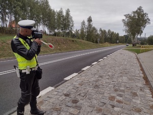 Zdjęcie kolorowe. Policjant ruchu drogowego stojąc na skraju drogi dokonuje pomiaru prędkości.