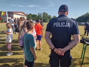 Zdjęcie kolorowe. Na pierwszym planie stoi odwrócony plecami policjant, z jego lewej strony stoi chłopiec w zielonej koszulce. W tle widać inne dzieci