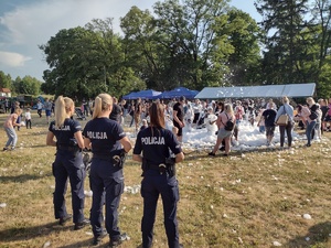 Zdjęcie kolorowe. Trzy policjantki odwrócone plecami stoją na tle dzieci bawiących się w pianie